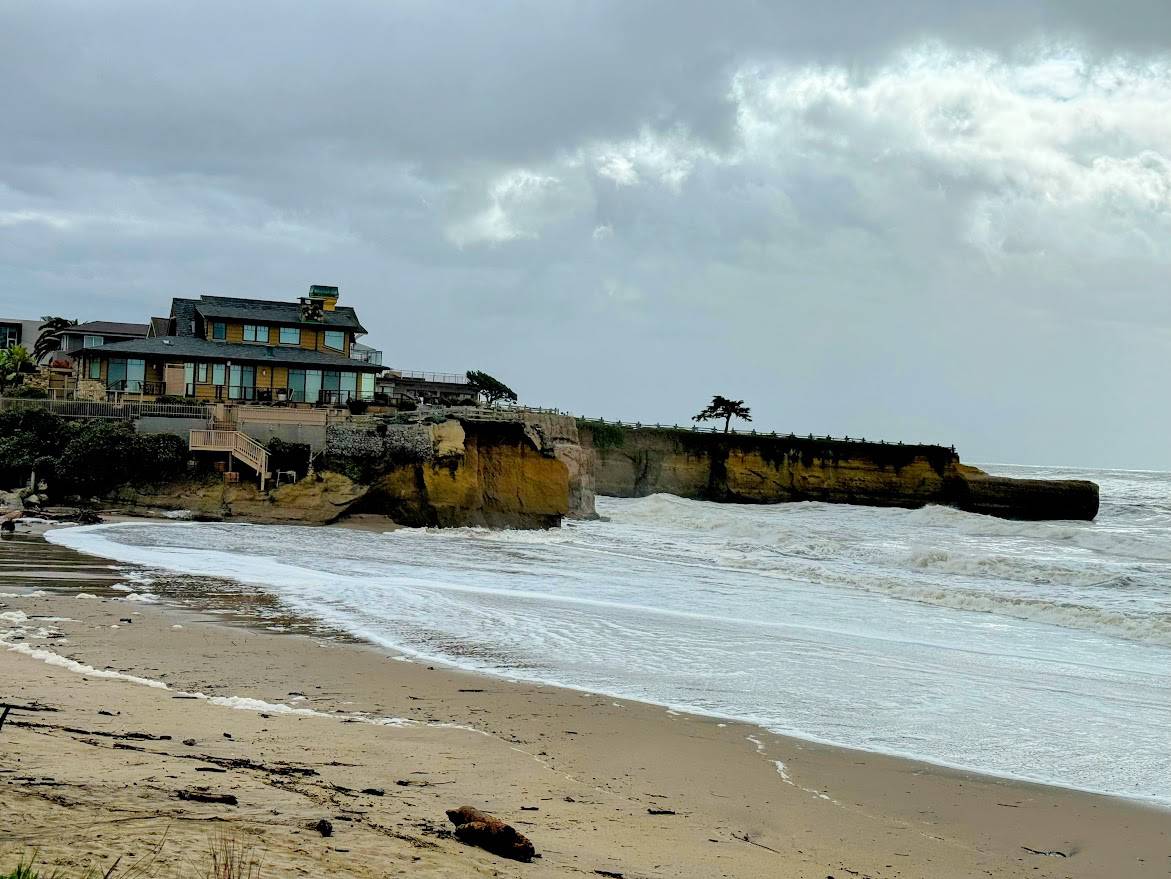 A beach with gentle waves washing onto the sand, backed by a rugged cliff with a modern house perched on top. A single windswept tree stands at the edge of the cliff, and the sky is overcast with thick clouds.