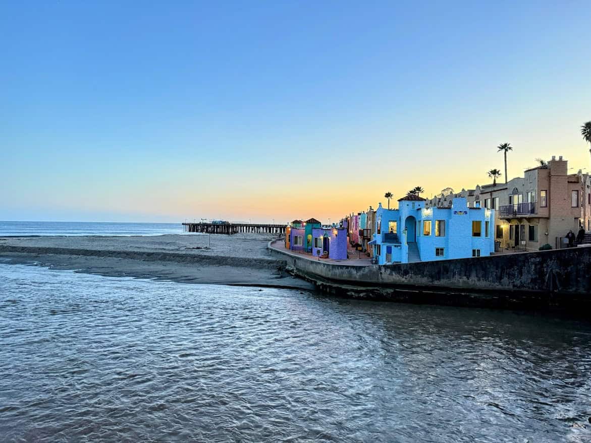 A scenic sunset at Capitola Beach, with pastel-colored Mediterranean-style buildings lining the waterfront and a wooden pier stretching into the calm ocean.