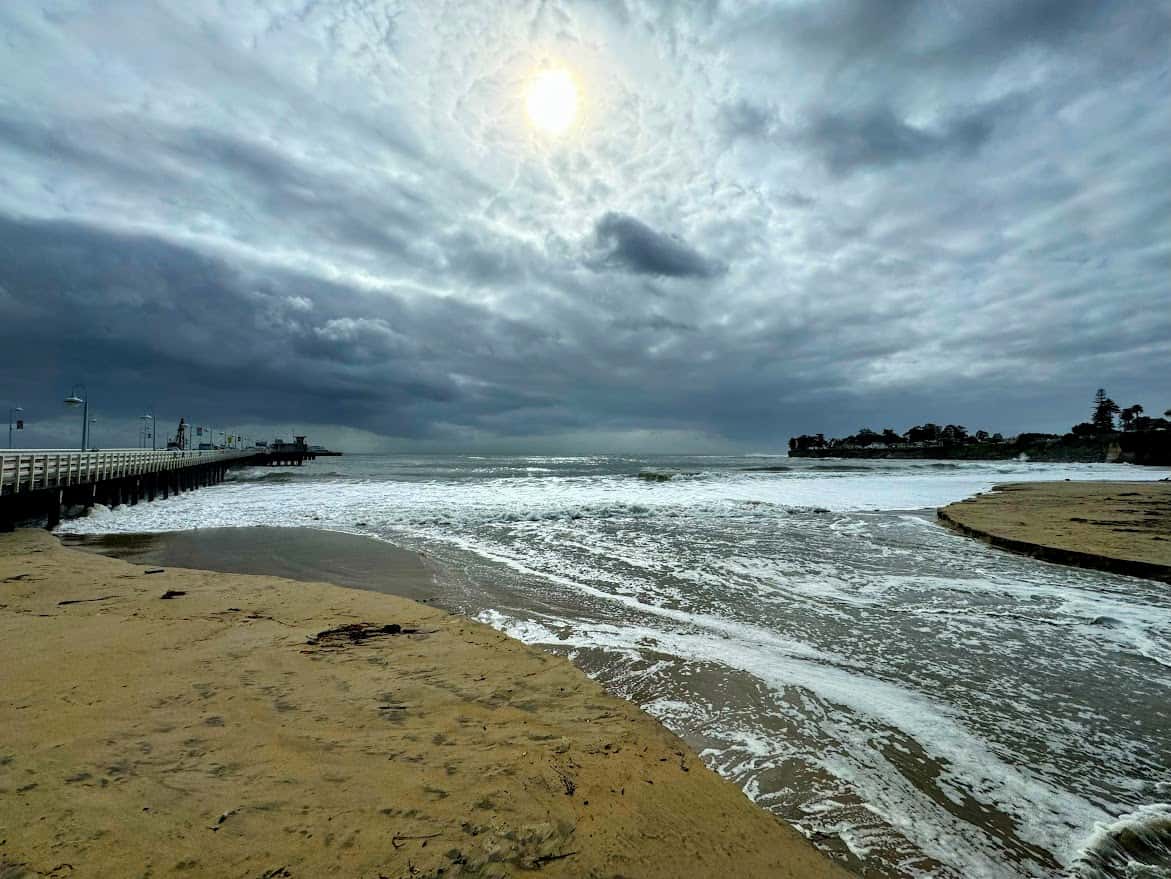 A dramatic scene at Cowell Beach, with stormy clouds above, waves rolling onto the shore, and the wooden Santa Cruz Wharf extending into the ocean.