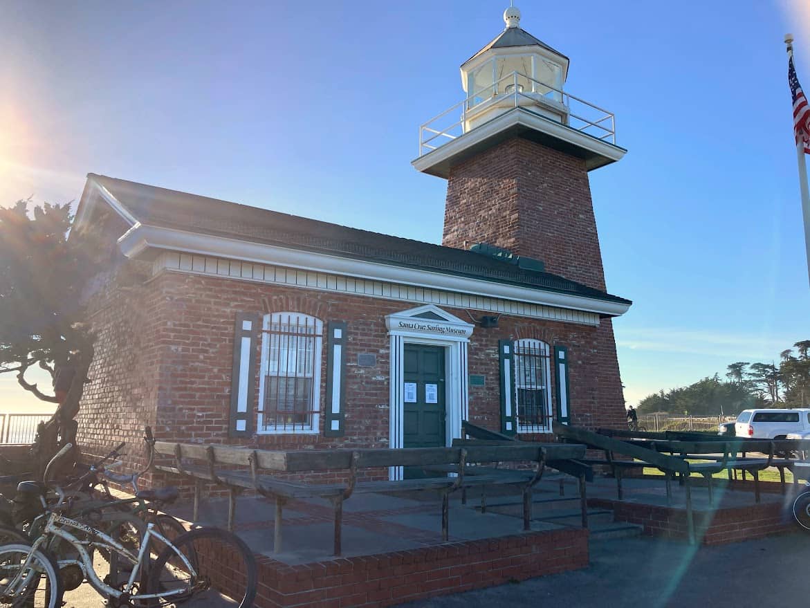 A historic red-brick lighthouse with a white lantern tower stands under a clear blue sky. Bicycles are parked nearby, and an American flag waves in the breeze.