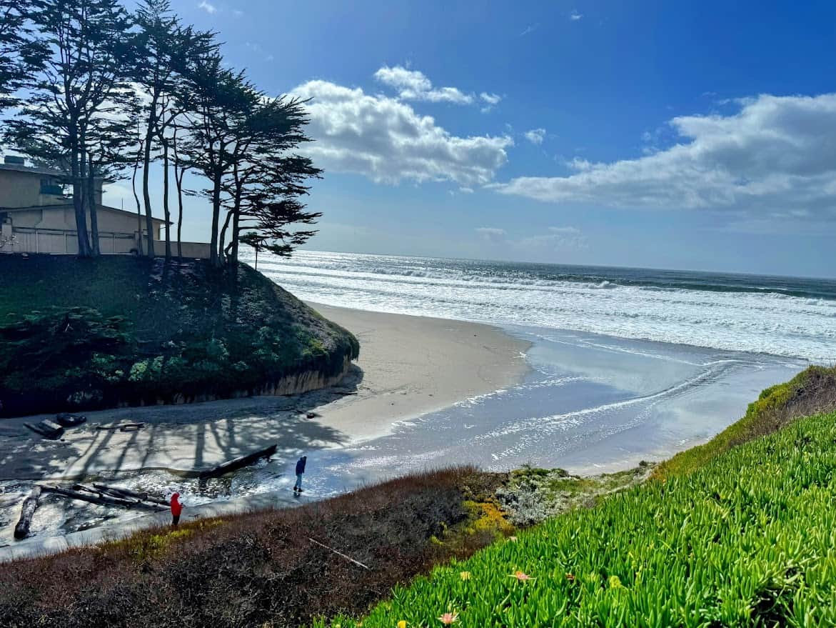 A peaceful cove at Manresa Main State Beach, with two people walking near the water, waves rolling onto the shore, and a house nestled among the trees.