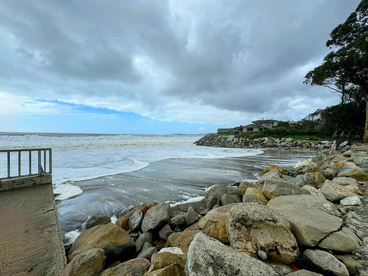 A rocky shoreline with ocean waves washing over a wet concrete ramp, leading towards a beachfront house with lush greenery. The sky is overcast with dark clouds, and a line of trees stands on the right side.