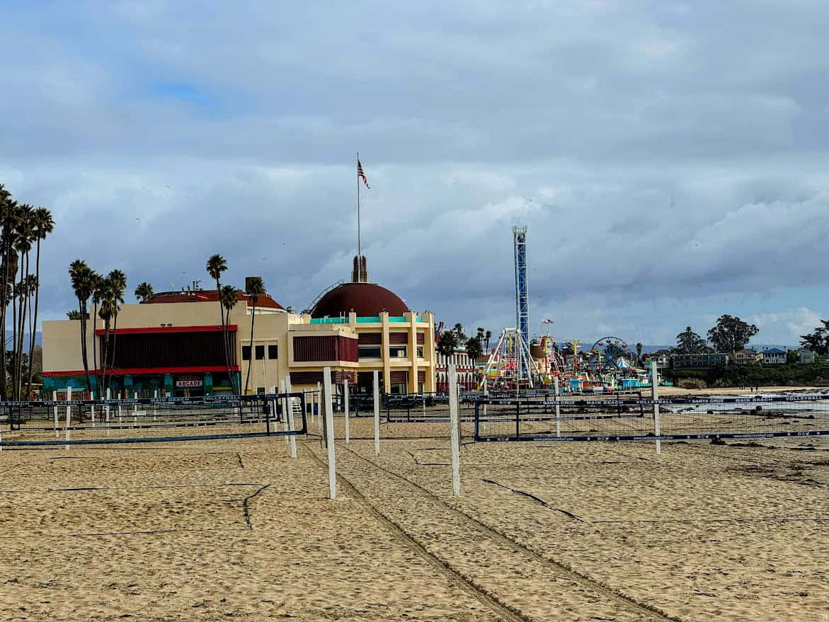 A wide view of Santa Cruz Main Beach with volleyball nets on the sand, and the historic Santa Cruz Beach Boardwalk amusement park in the background.