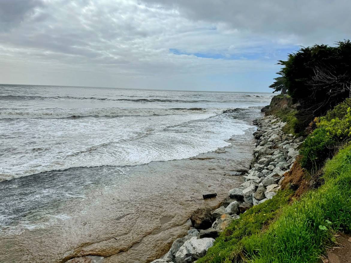 A rugged shoreline with jagged rocks and green coastal vegetation lining the edge of the land. Powerful ocean waves roll towards the shore under a cloudy sky.