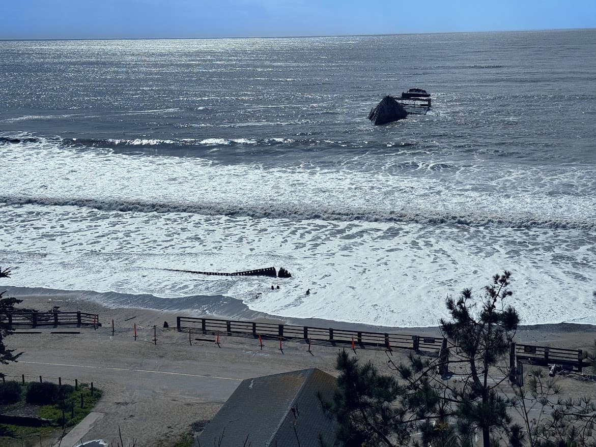 A view of Seacliff State Beach in California, featuring the partially sunken remains of the SS Palo Alto, a concrete ship off the coast. White-capped waves roll onto the sandy shore, while a wooden fence lines the beach. The bright blue sky reflects on the shimmering ocean surface.
