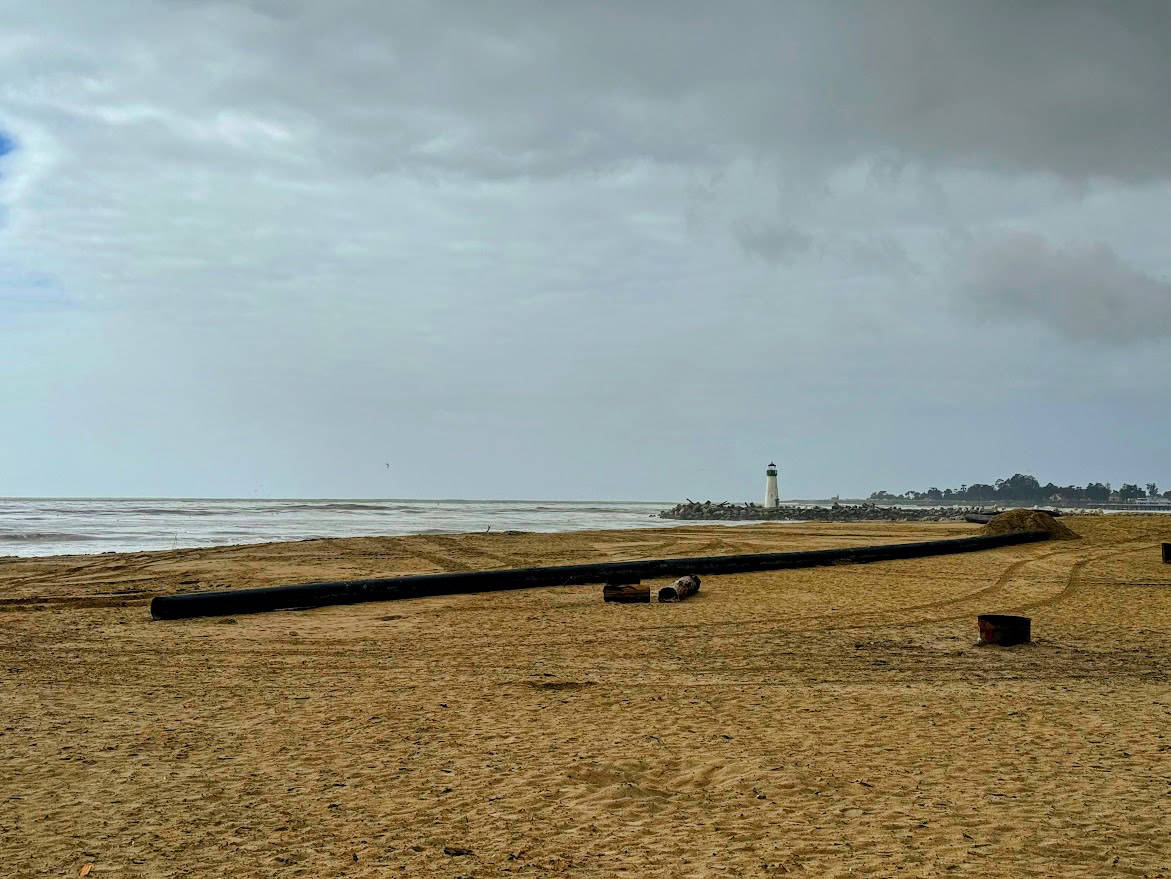 A view of Twin Lakes State Beach with a long black pipe on the sand leading toward the distant Walton Lighthouse under a gray sky.