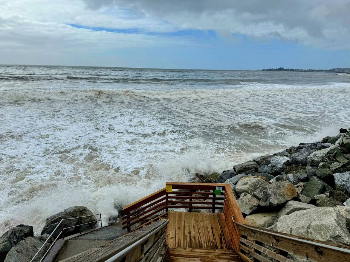 A wooden staircase descends towards the ocean as large waves crash against the rocky shore. The horizon stretches under a partly cloudy sky, and a distant coastline is visible in the background.