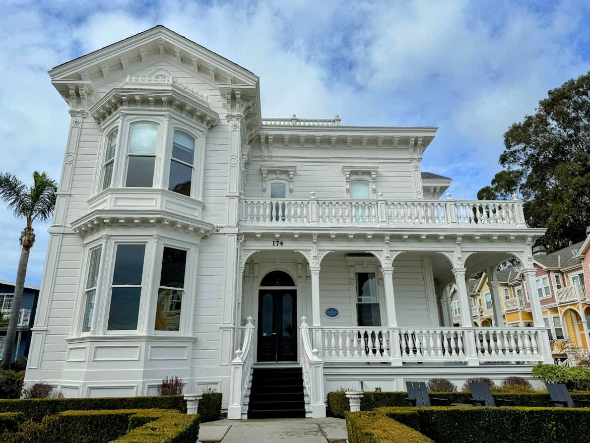 A grand white Victorian-style house with intricate balconies and bay windows. Palm trees and neatly trimmed hedges surround the home, and pastel-colored houses line the street.