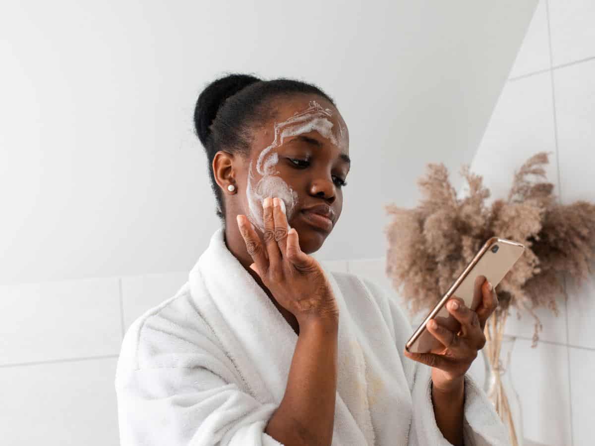 A woman applying facial cleanser while looking at her phone, highlighting a necessary skincare item in the California Packing List for maintaining clear skin during travel.