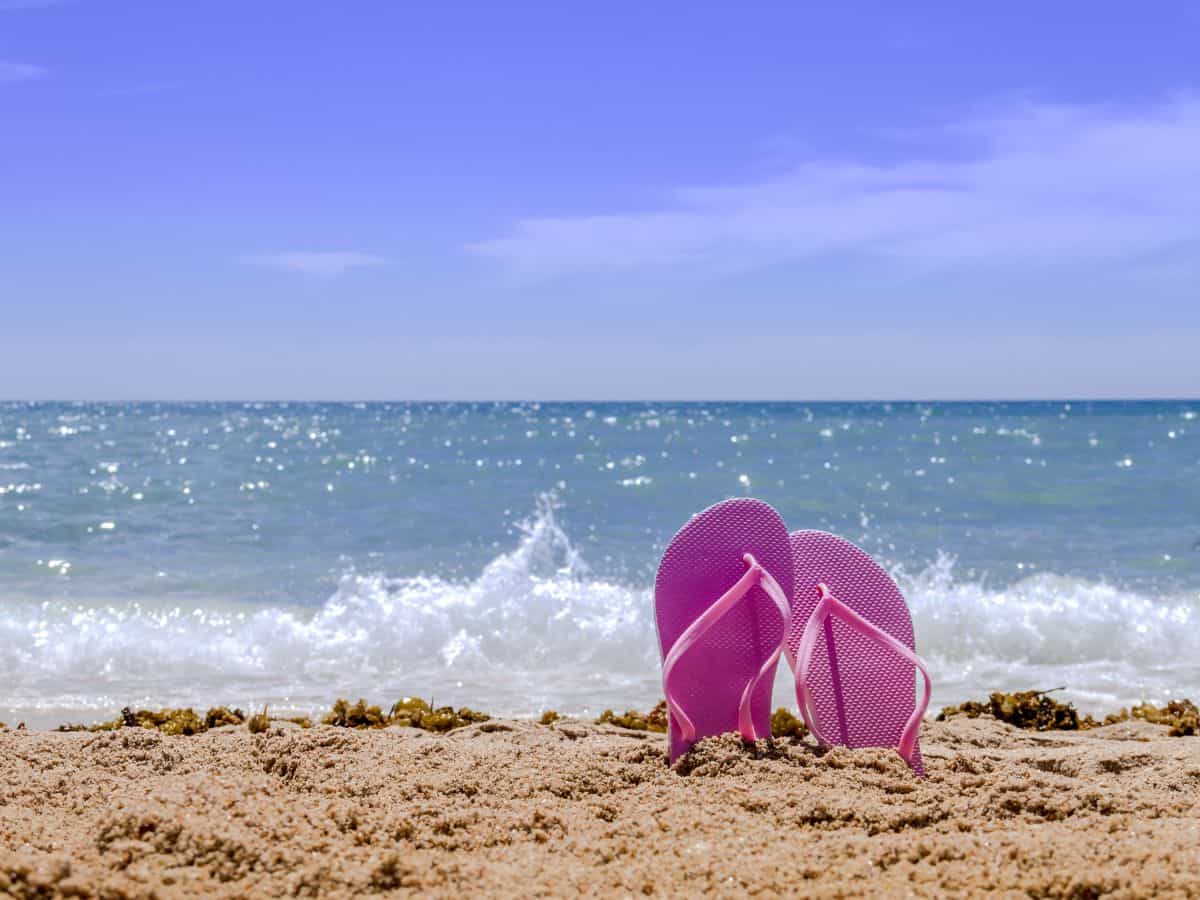 A pair of pink flip flops placed on a sandy beach with waves gently rolling in the background. Flip flops are a must-have item on your California packing list for enjoying the beautiful beaches.