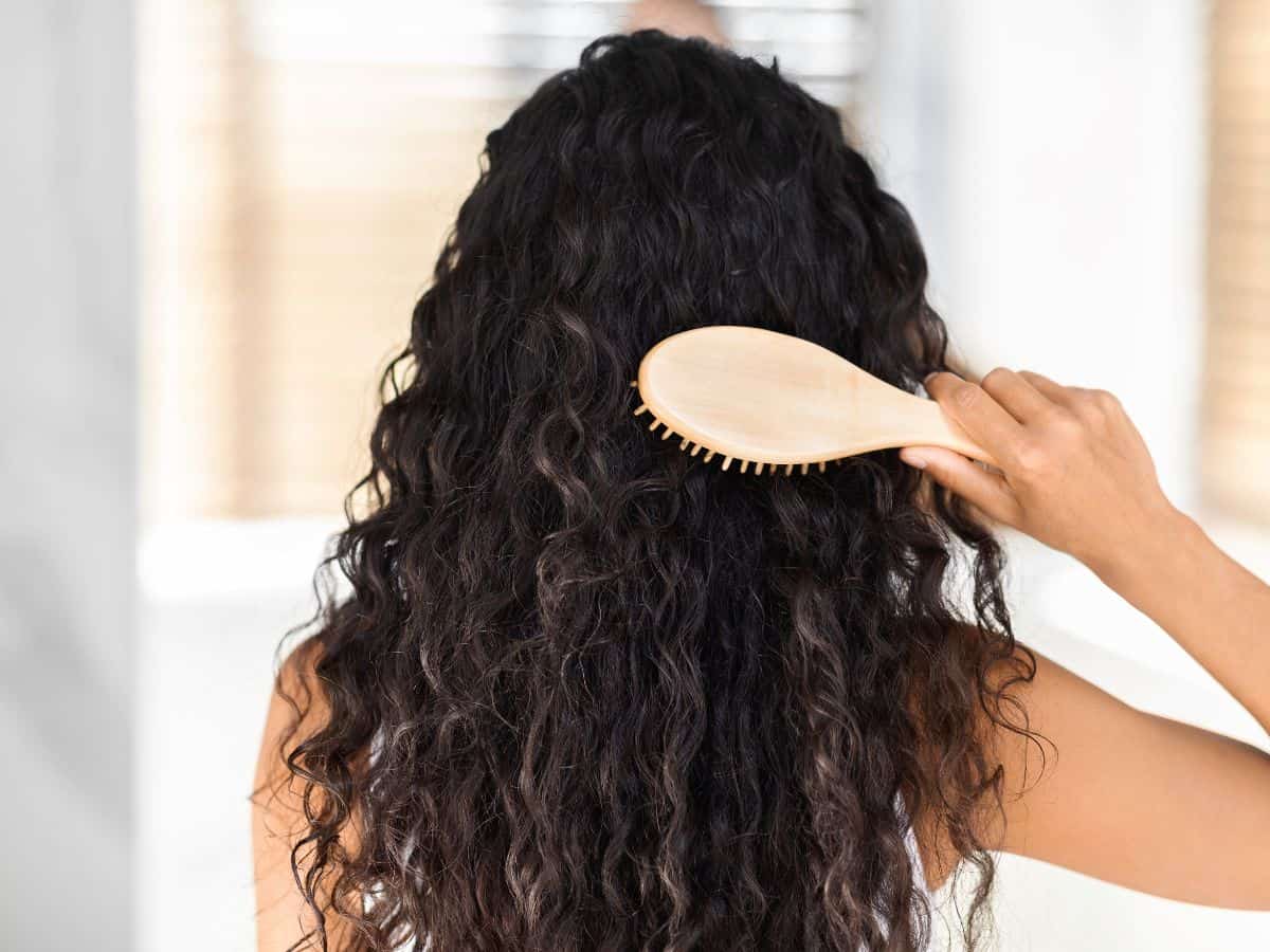 A woman with long, curly hair brushing her hair with a wooden brush. A must-have for the California Packing List to manage hair in the coastal breeze.