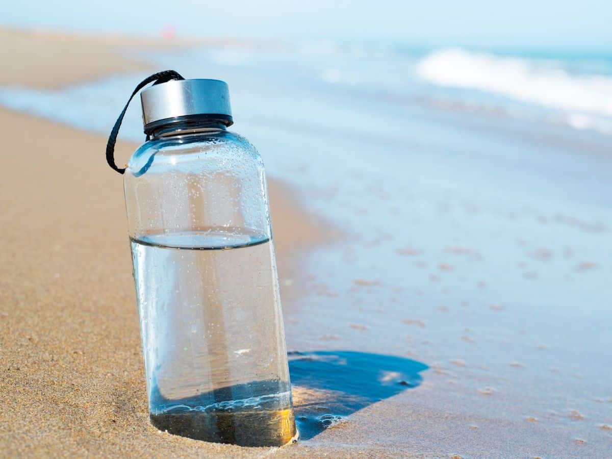 A reusable water bottle resting on a sandy beach with the ocean in the background. A reusable water bottle is an eco-friendly item to add to your California packing list to stay hydrated.