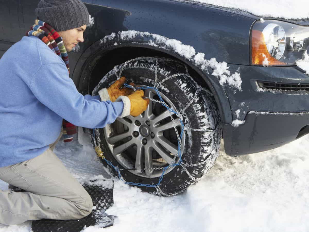 A person installing snow chains on a car tire in a snowy environment. Snow chains are a necessary addition to your California packing list if traveling to mountainous regions during winter.