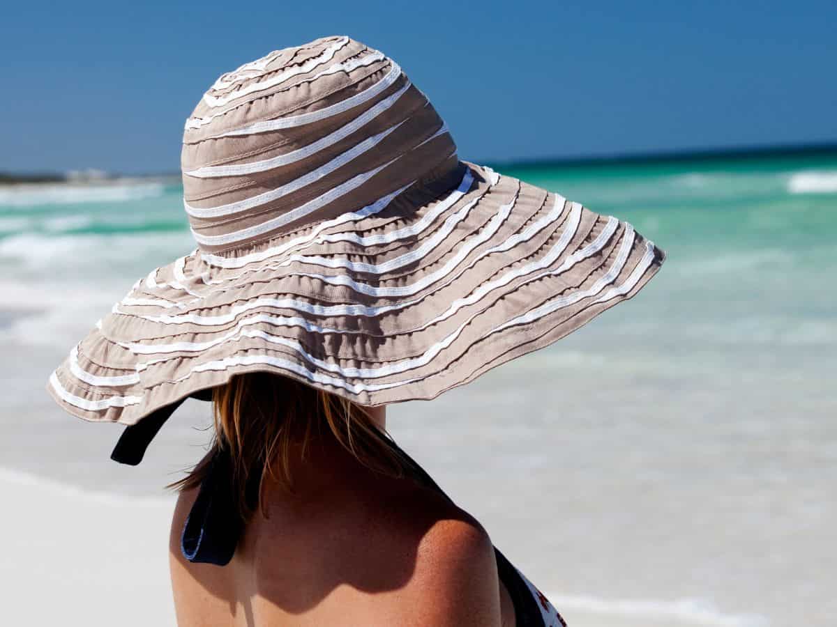 A woman wearing a large, beige striped sunhat on a sunny California beach, gazing at the turquoise ocean in the background. Essential for the California Packing List to protect from the sun.

