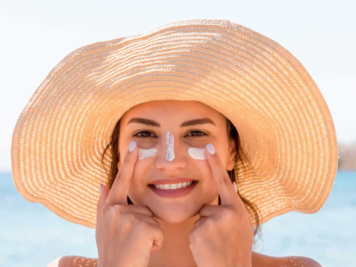  A woman applying sunscreen to her face, wearing a wide-brimmed hat on a sunny beach. Sunscreen is a critical item on your California packing list to protect your skin from harmful UV rays.