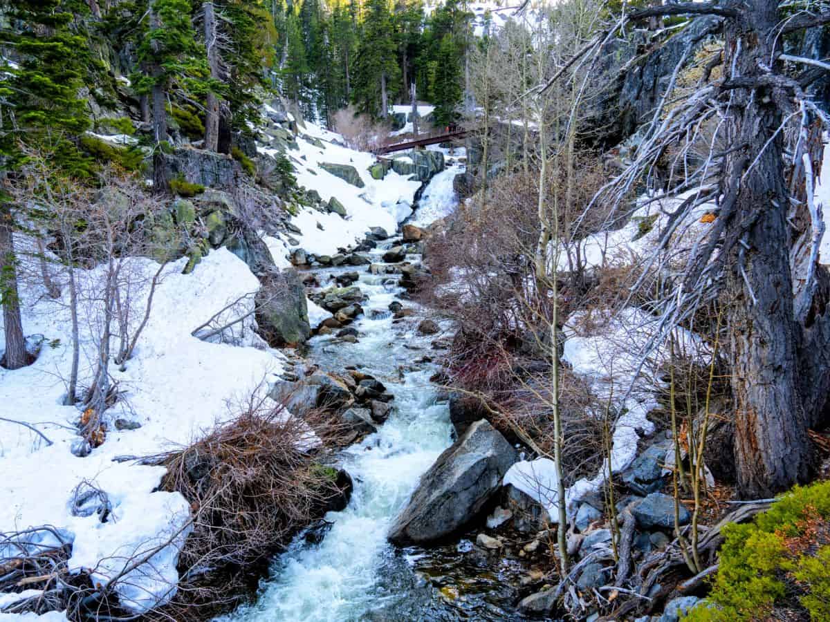 The rushing waters of Eagle Falls cascade through a snowy landscape, surrounded by rocky terrain and dense pine trees. This trail is a picturesque and easy hike, making it one of the 11 Easy Lake Tahoe Hikes To Do for those who love waterfalls and winter scenery.
