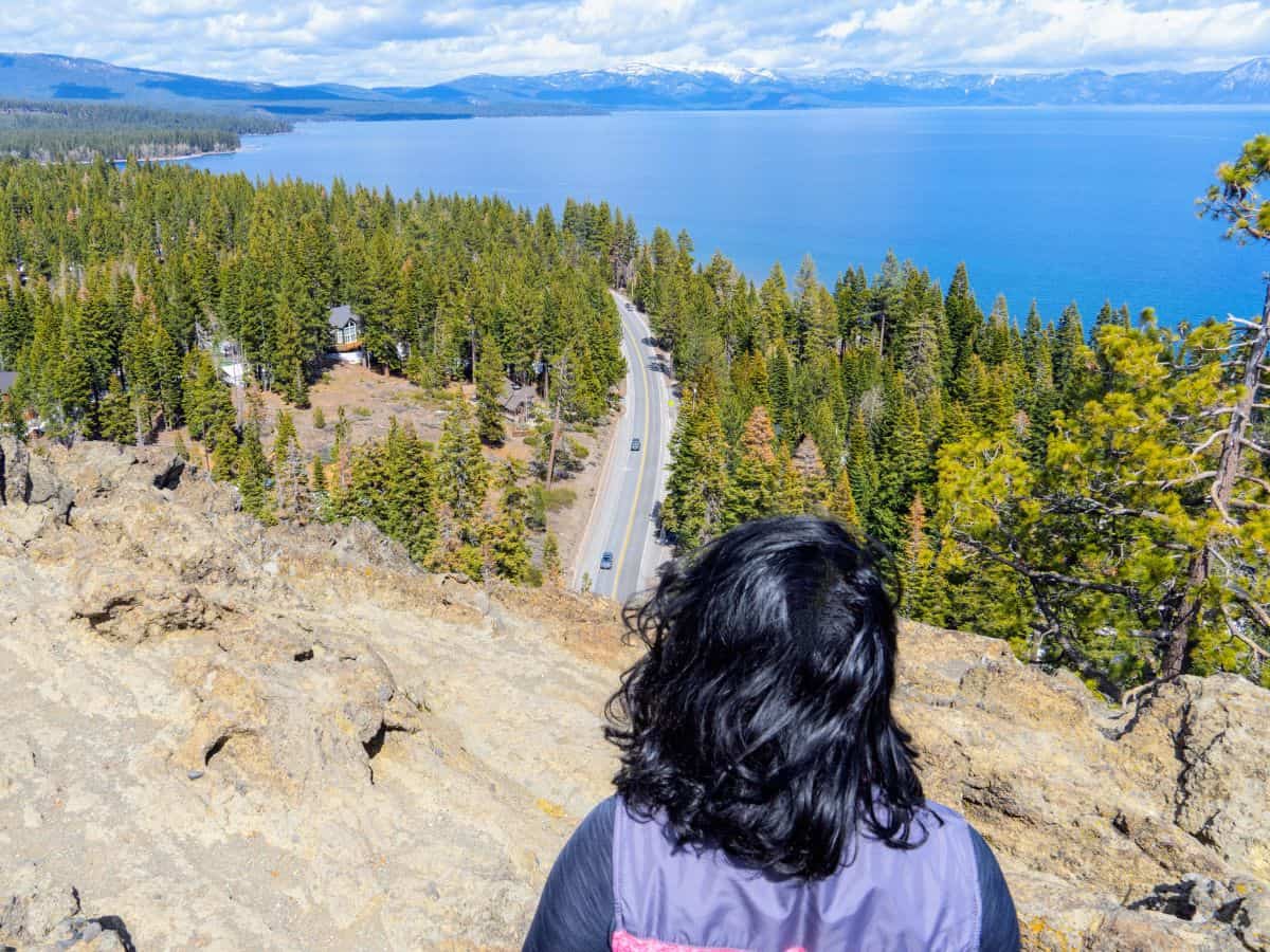 A hiker overlooks Lake Tahoe from the top of Eagle Rock Trail, with a winding road and dense forest below. This easy trail provides rewarding views, making it one of the 11 Easy Lake Tahoe Hikes To Do for a quick yet scenic outdoor experience.