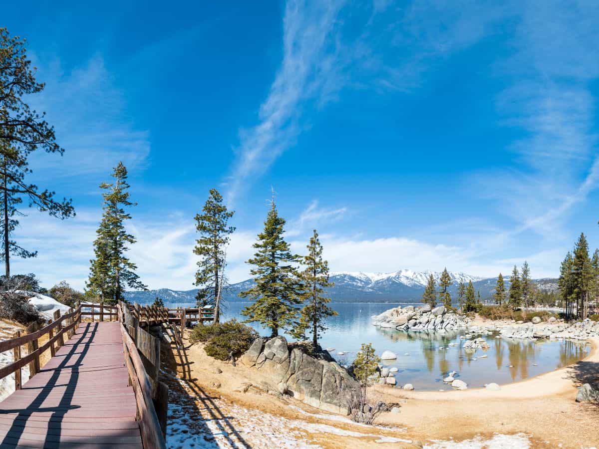 The boardwalk of Sand Harbor Nature Trail leads through a picturesque landscape of pine trees and rocky shorelines, with Lake Tahoe's pristine waters and snow-capped mountains in the background. This easy and accessible trail is one of the 11 Easy Lake Tahoe Hikes To Do.