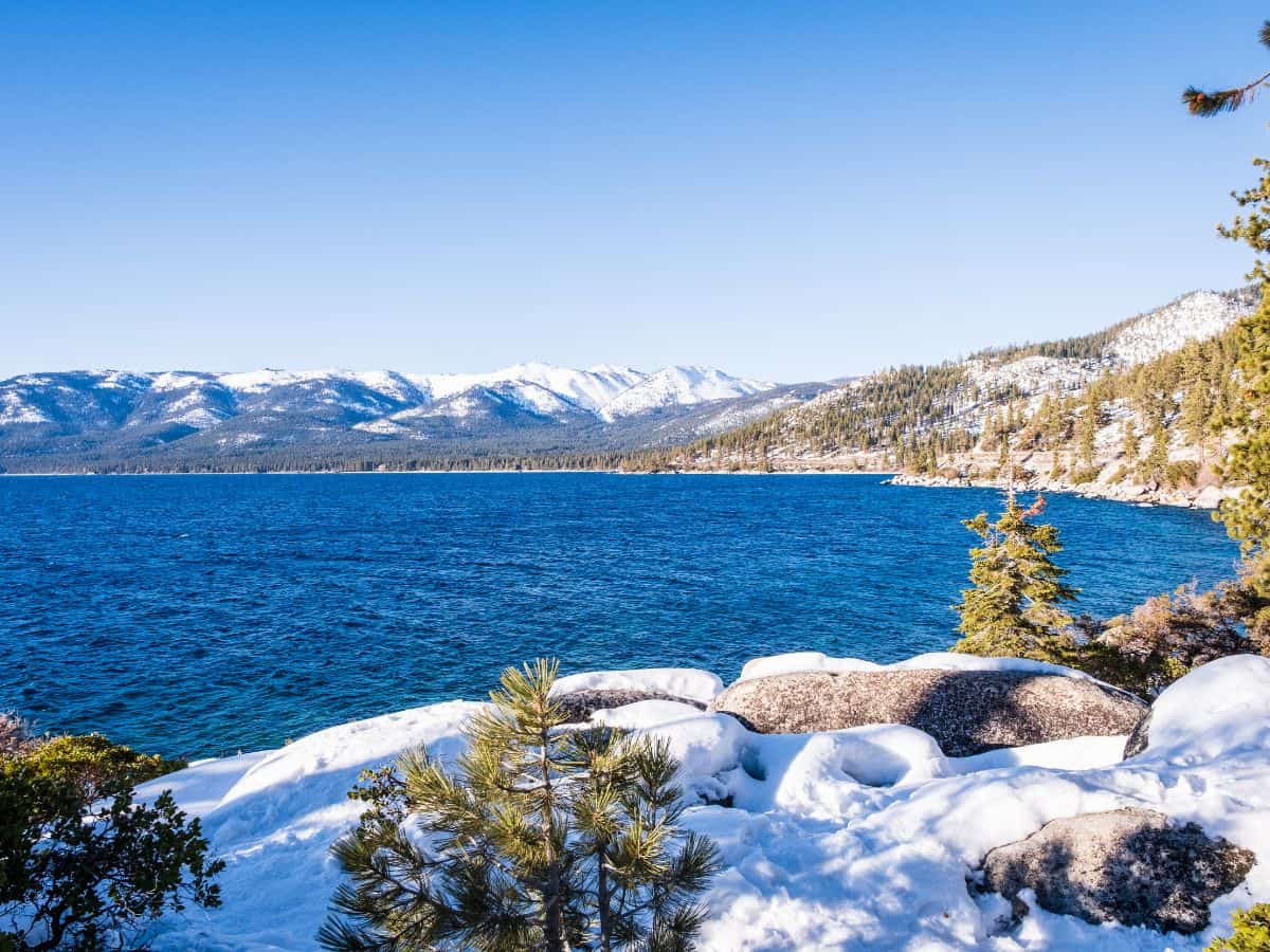 A snow-dusted view along the Tahoe East Shore Trail, where the clear blue waters of Lake Tahoe meet the snowy mountains in the background. This trail offers easy access to stunning winter scenes, making it one of the 11 Easy Lake Tahoe Hikes To Do.