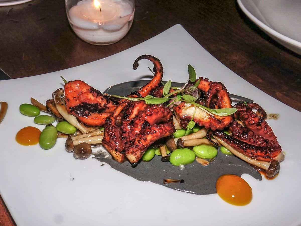 Char-grilled octopus served with edamame and mushrooms on a modern plate, featuring vibrant colors and textures, epitomizing the creative approach to Best Seafood In Southern California. The grilled octopus adds a smoky touch to this innovative seafood dish.