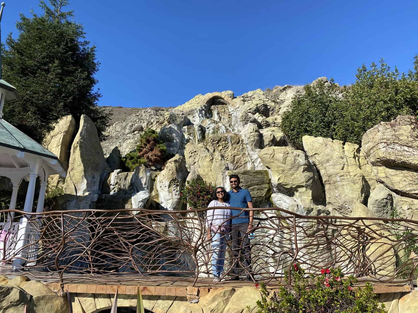 Shreeyeh and Kiran standing on a decorative bridge with an intricate, nature-inspired railing. Behind them is a rocky landscape featuring a small waterfall and surrounded by greenery, under a bright blue sky. A gazebo with a green roof is partially visible to the left.