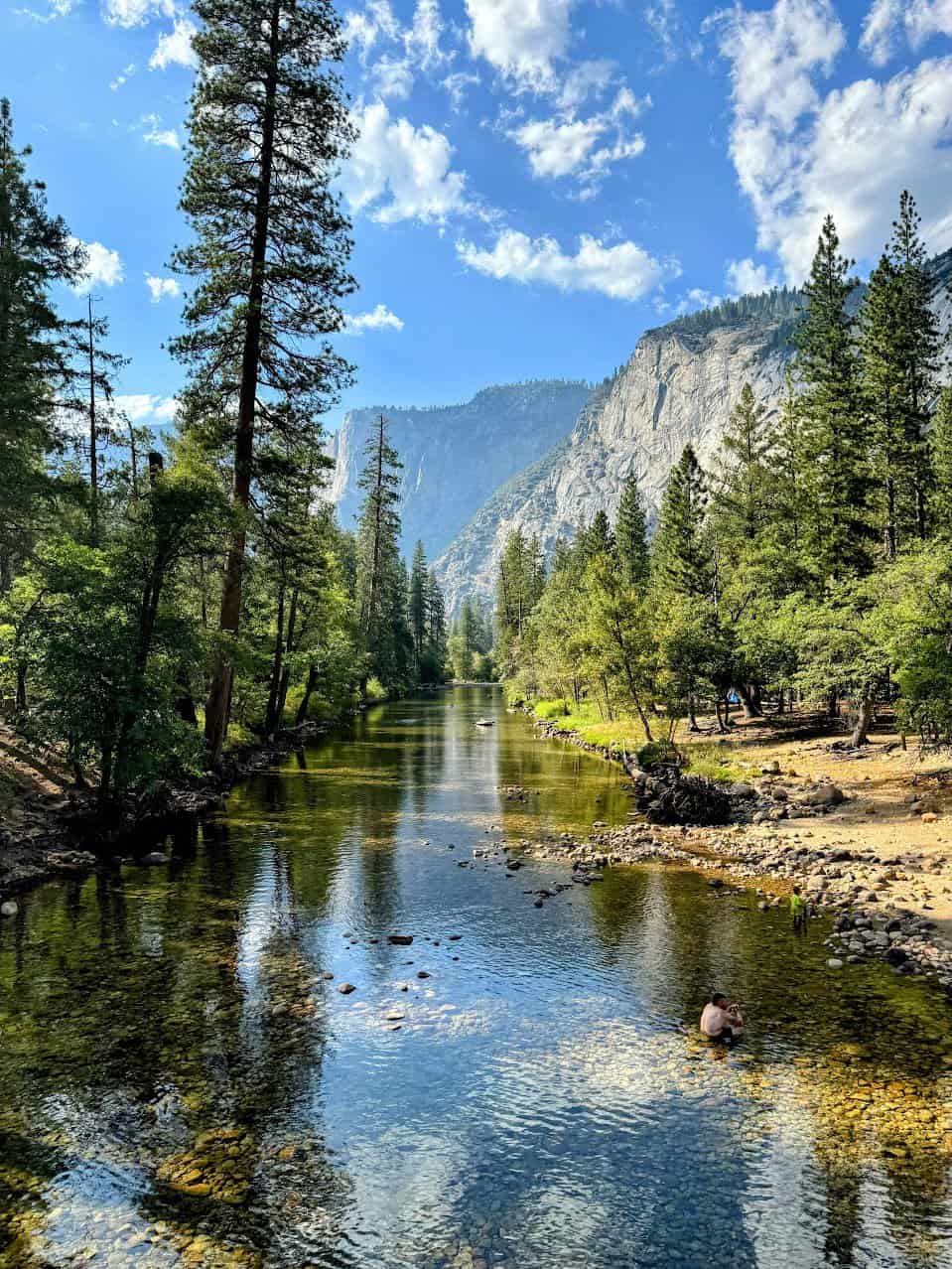 A serene view of the Merced River flowing gently through Yosemite National Park, surrounded by towering pine trees and rugged granite cliffs in the background. The clear water reflects the blue sky and scattered clouds, with a person sitting in the shallow riverbed adding a peaceful touch to the scene.
