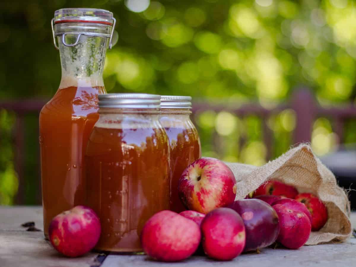 Glass bottles filled with freshly pressed apple cider sitting beside a basket of apples. Apple cider tasting is one of the flavorful things to do in California in October, especially for visitors exploring the apple orchards and harvest festivals.
