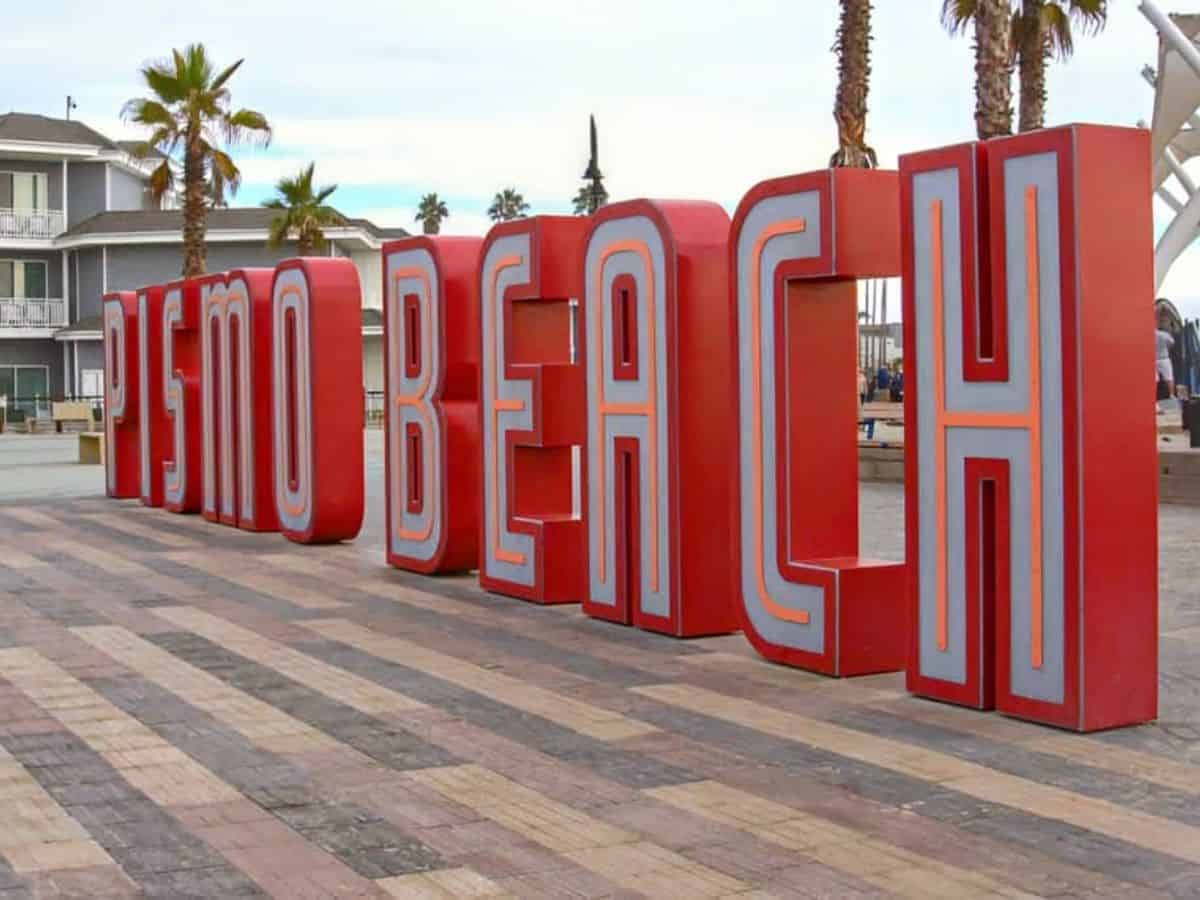 A large red sign spelling out "Pismo Beach" stands prominently on a boardwalk near the ocean. Visiting the Central Coast beaches is one of the refreshing things to do in California in October, providing a beautiful coastal escape with mild autumn weather.