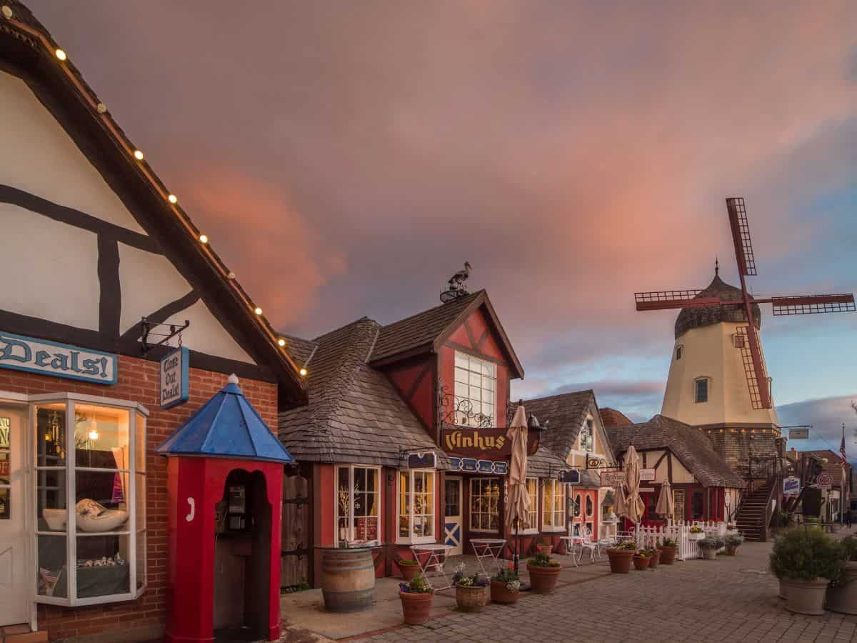 Charming, colorful buildings with a windmill in the background at sunset, creating a fairytale-like scene in the Danish village of Solvang. Stepping into this picturesque town is one of the magical things to do in California in October, with cozy shops and a warm autumn ambiance.