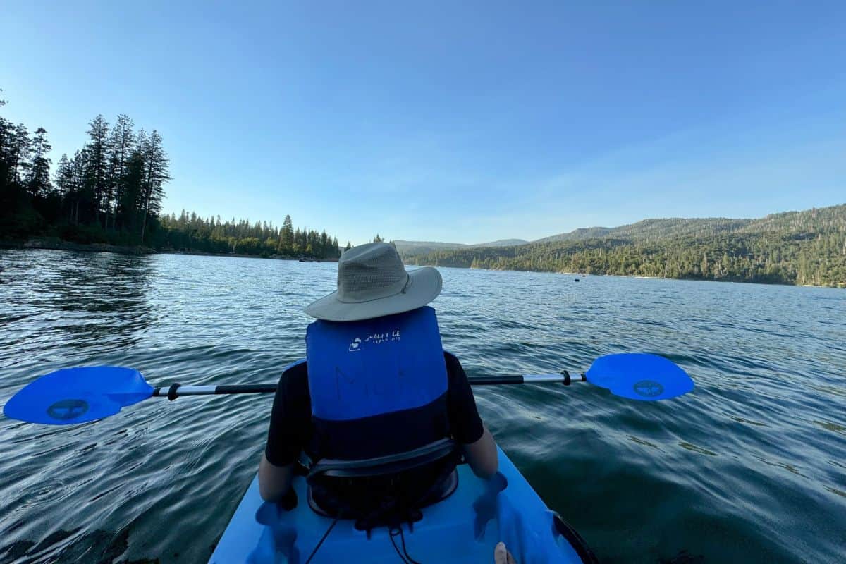 A peaceful scene on Bass Lake, California, with a kayaker paddling through calm waters surrounded by forested hills under a clear blue sky. This serene location offers outdoor adventure and relaxation on the Los Angeles to Yosemite road trip.