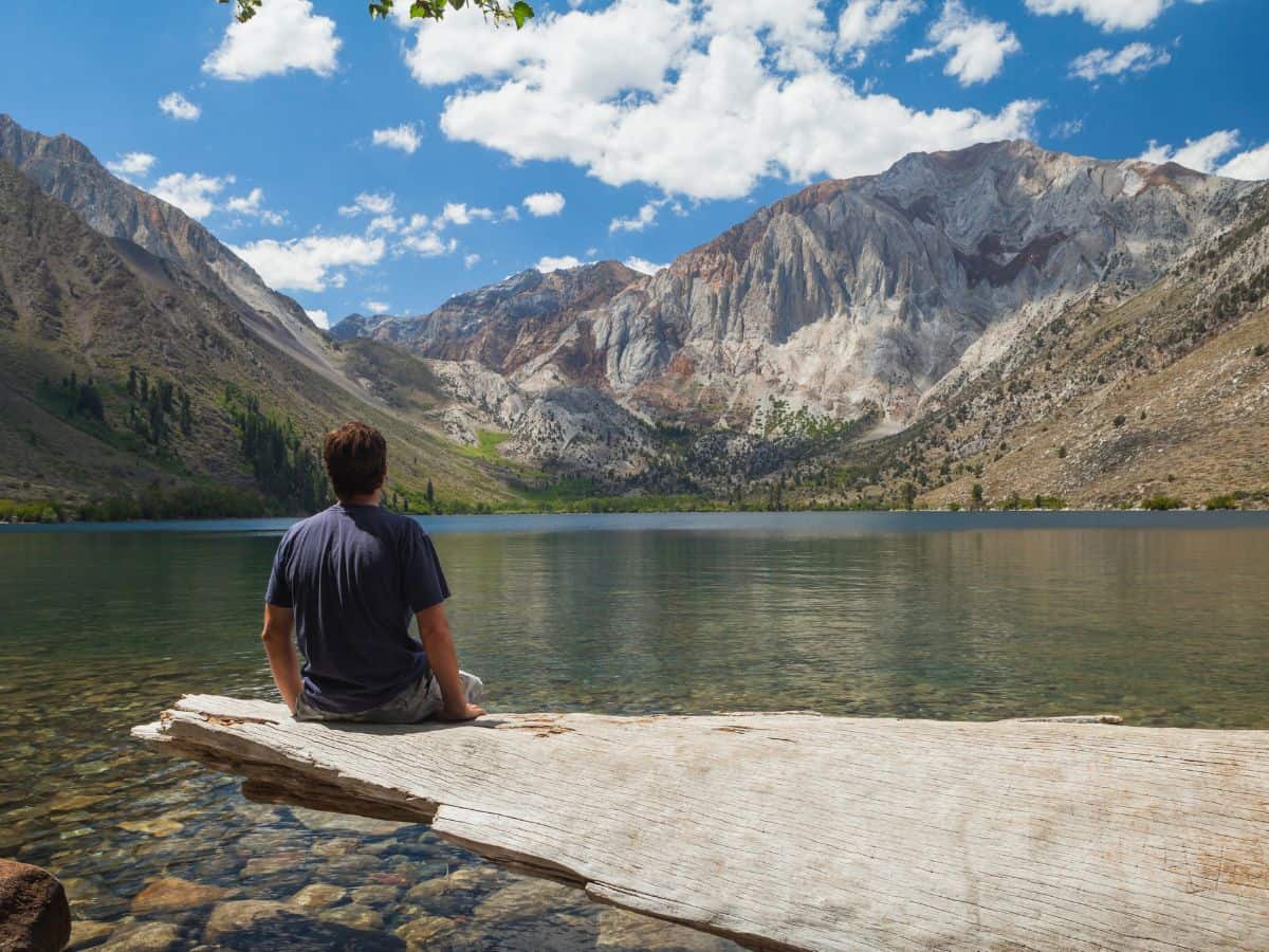 A picturesque view of Convict Lake as part of a Death Valley to Yosemite road trip. The image features a man sitting on a large log near the crystal-clear lake, gazing at the majestic mountains towering over the water. The mountains, with their steep cliffs and patches of greenery, are set against a vibrant blue sky with white clouds.