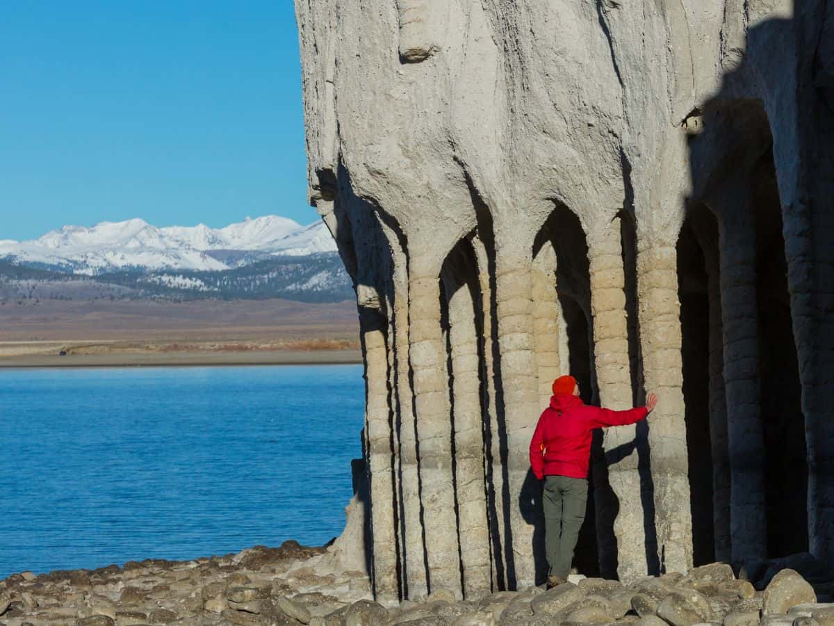 A traveler standing at the iconic columns of Crowley Lake on a Death Valley to Yosemite road trip. The individual, dressed in a red jacket, touches one of the large stone pillars carved out by nature. Behind them, the still blue waters of Crowley Lake stretch out, with snowy mountains visible in the distance, offering a scenic contrast between stone, water, and sky.