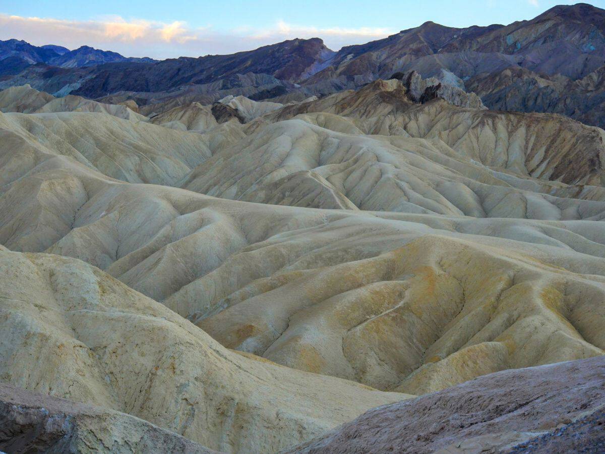 A stunning view of Death Valley National Park, featuring vast golden and beige sand dunes with soft, curving shapes. The barren, desert landscape extends into the background with rugged, multi-colored mountains lining the horizon. The sky above is mostly clear with a touch of clouds, reflecting the vast and arid terrain encountered on a Death Valley to Yosemite road trip.