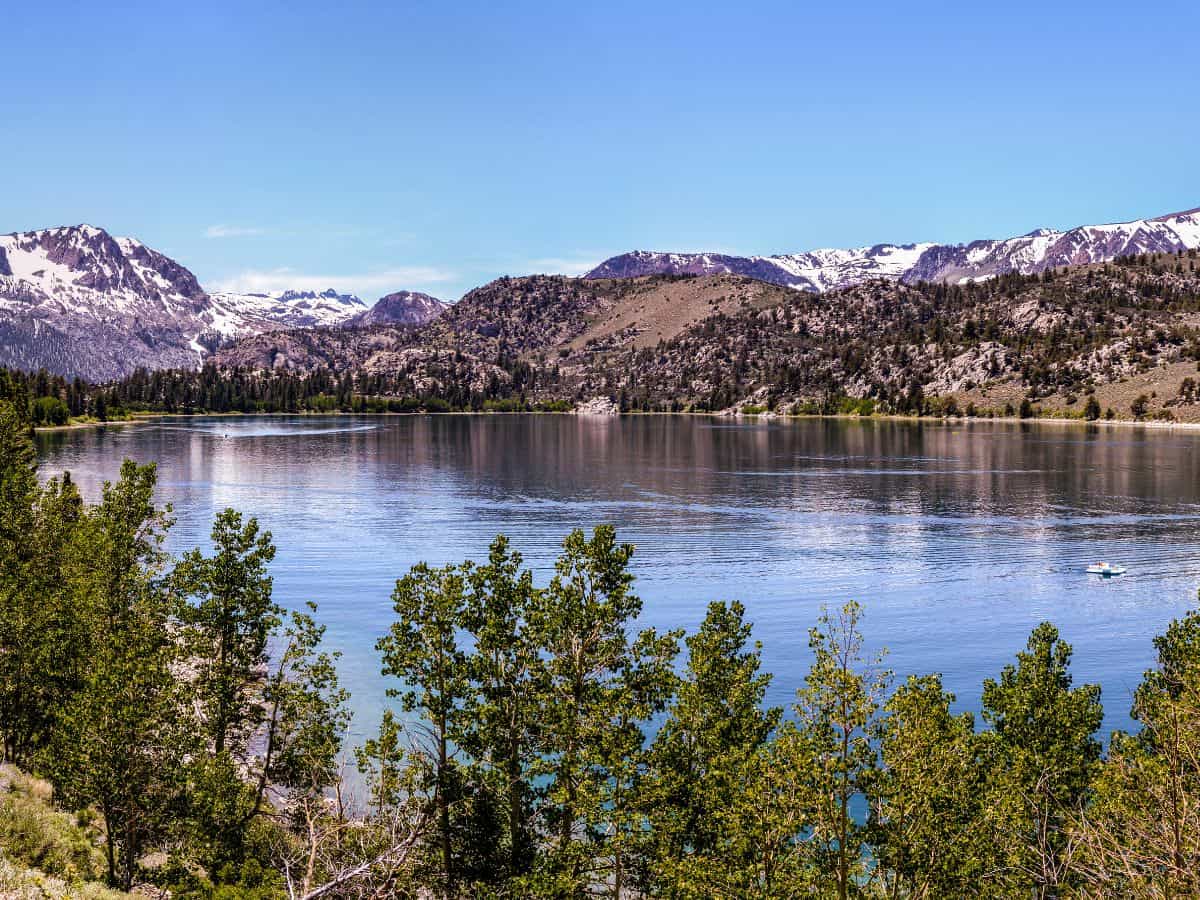 A stunning view of June Lake, framed by vibrant green trees along the shore, with a mirror-like surface reflecting the surrounding mountains. Snow-dusted peaks rise in the background, while the pristine blue water shimmers under the bright sky. The scenic vista is a highlight of the Death Valley to Yosemite road trip.