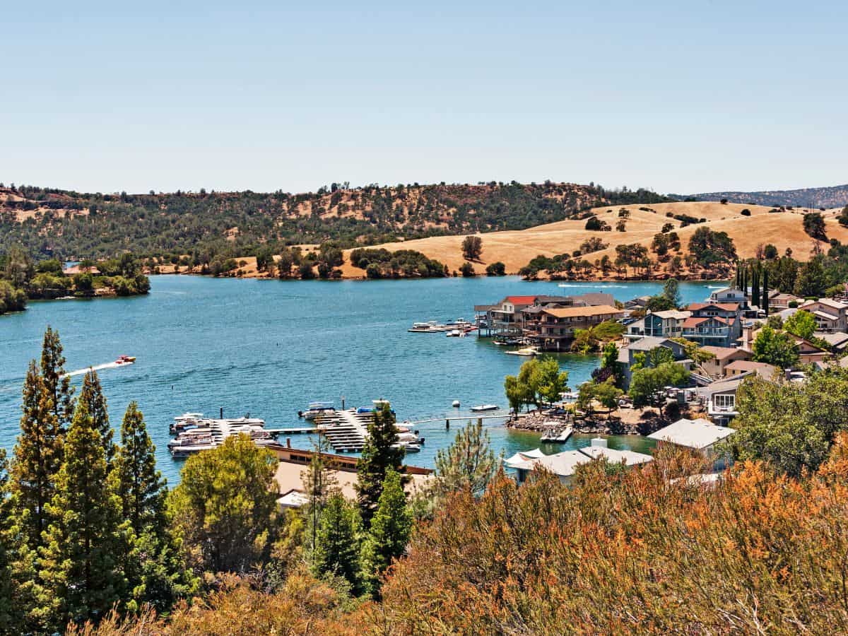 A serene view of Lake Tulloch in California, with boats docked near waterfront homes. Rolling hills and scattered trees surround the calm blue waters, creating a peaceful atmosphere during the Seattle to Yosemite road trip.