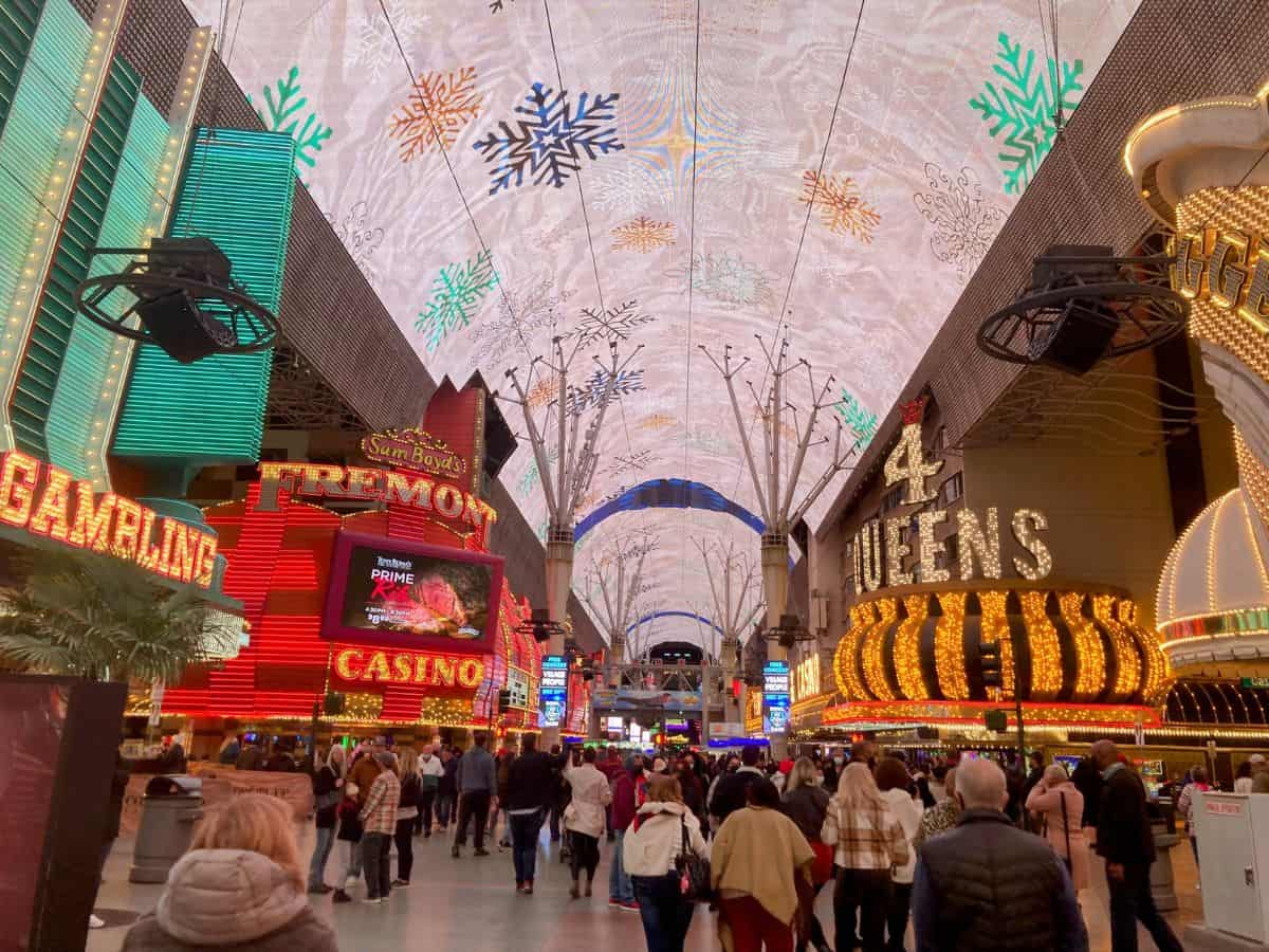 A vibrant scene from Fremont Street in Las Vegas, showcasing neon-lit casino signs like the iconic "Fremont" and "4 Queens" under a dazzling, colorful canopy of lights. The crowd-filled street is lively with visitors, capturing the bustling atmosphere of this famous stop on a Las Vegas to Yosemite Road Trip.