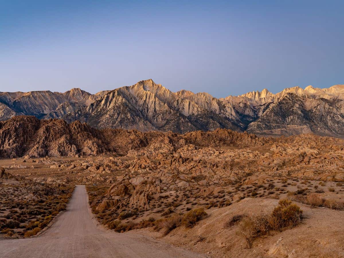 A serene nature scene in Bishop, California, located along the Death Valley to Yosemite road trip route. The image shows a peaceful river flowing through a forest with colorful autumn foliage. The trees reflect in the calm water, and the distant mountains peek through, creating a tranquil wilderness setting.
