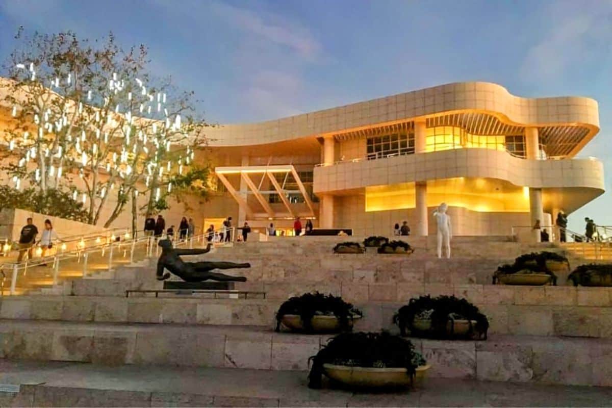 The Getty Center in Los Angeles illuminated by golden lights during dusk, with modern architecture and art sculptures in the foreground. Visitors walk up the steps toward the entrance, enjoying a serene evening. A perfect stop on a road trip from Los Angeles to Yosemite, showcasing a blend of art and culture.