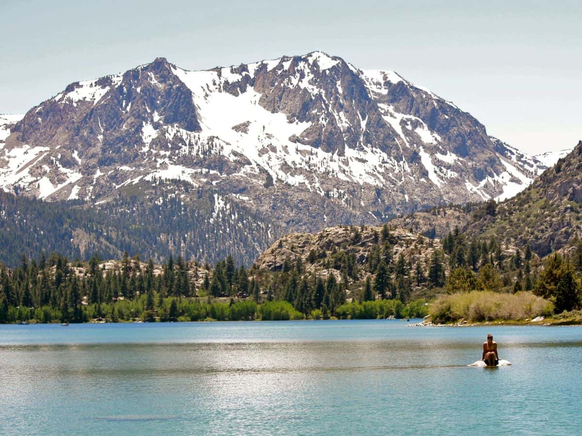 A tranquil scene from Mammoth Lakes on a Death Valley to Yosemite road trip. The photo shows a lake surrounded by forest, with snow-capped mountains in the background. The shimmering water reflects the surrounding trees and mountains, while a single figure paddles across the lake, adding a peaceful and adventurous element to the scene.