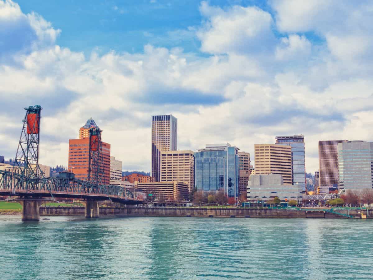 : The skyline of Portland, Oregon, with its mix of modern and historic buildings, set against a partly cloudy sky. The image features the Willamette River in the foreground and a bridge connecting the city. Taken during a Seattle-to-Yosemite road trip, this scene captures the essence of Portland.