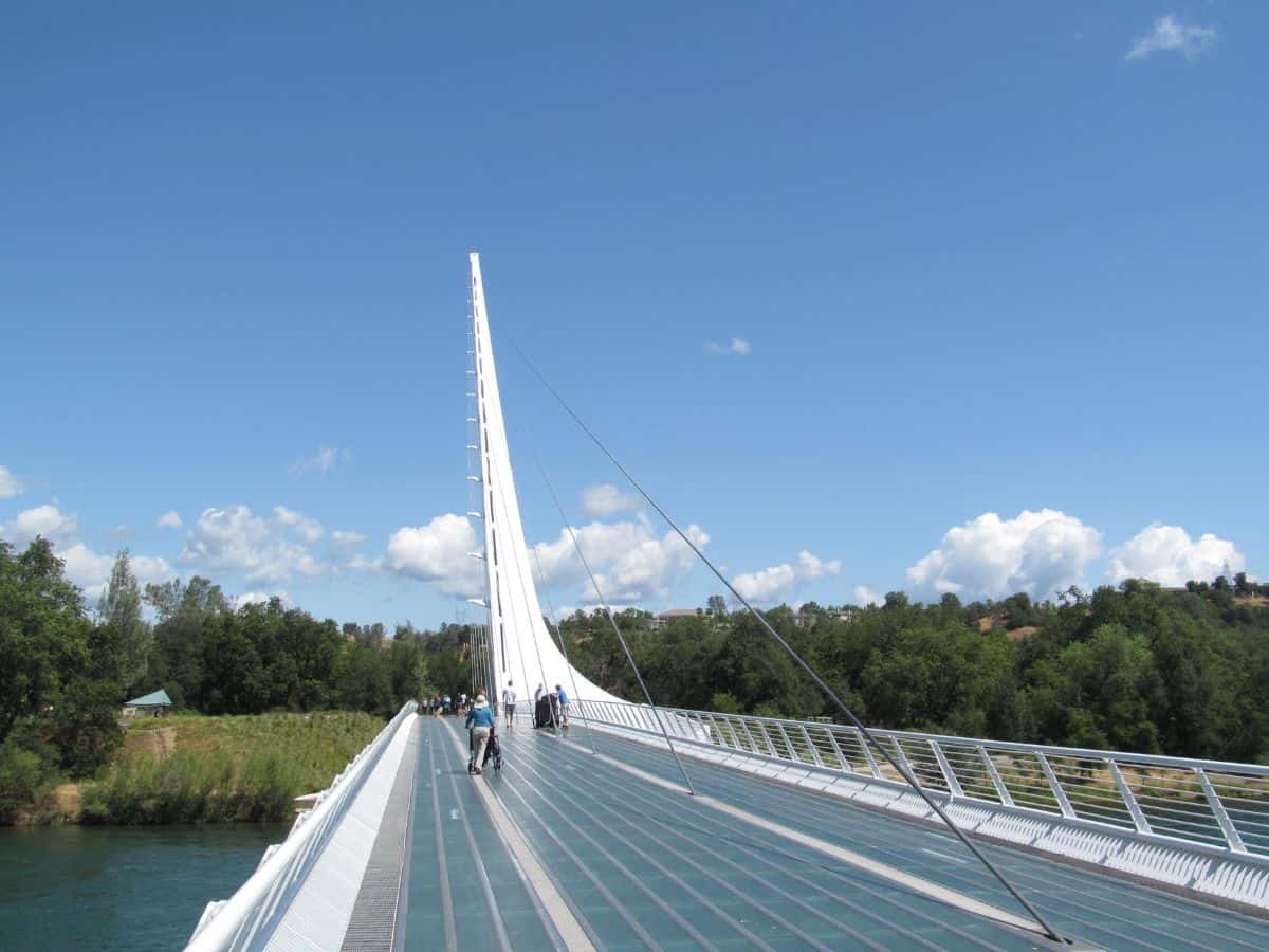 A view of the Sundial Bridge in Redding, California, on a sunny day. The sleek, white architectural structure contrasts with the blue sky and lush greenery on either side of the river. A scenic stop during the Seattle-to-Yosemite road trip.