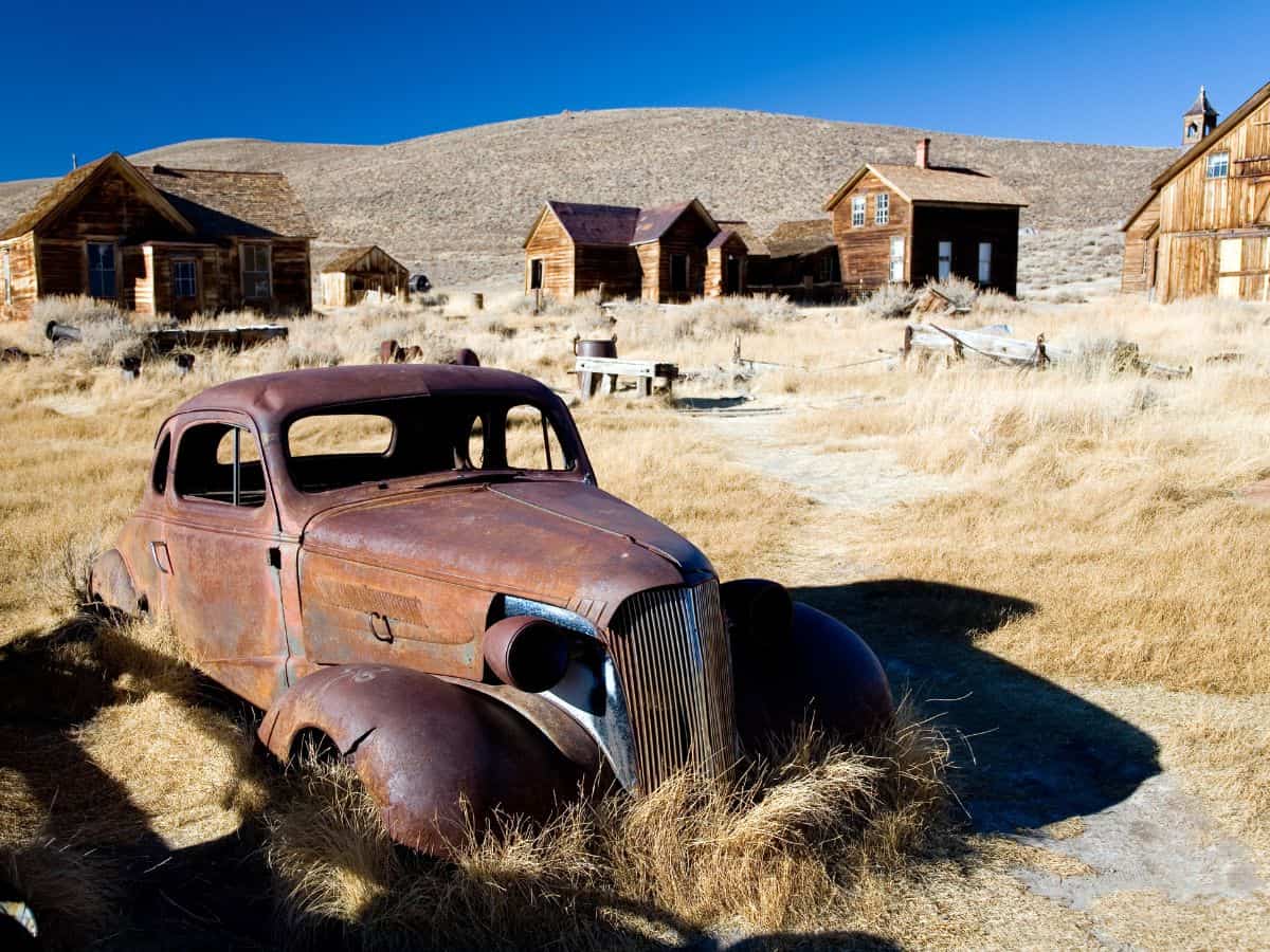 A haunting image of an old, rusted car sitting in the ghost town of Rhyolite, Nevada. Surrounded by abandoned wooden buildings, the scene evokes the forgotten past of this once-thriving mining town, now a fascinating stop on a Las Vegas to Yosemite Road Trip for those interested in exploring ghost towns.