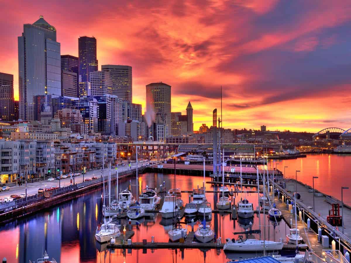 A vibrant sunset over the Seattle skyline as seen from the waterfront, with skyscrapers reflecting orange, pink, and purple hues in the sky. Sailboats are docked at the marina, and the cityscape includes iconic tall buildings. This is a beautiful view captured during a Seattle to Yosemite road trip.