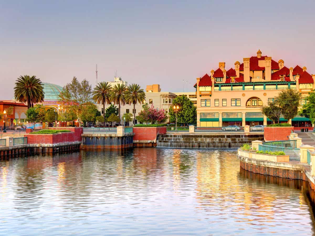 A picturesque view of downtown Stockton, California, taken from the waterfront. Palm trees line the canal, and a historic red-roofed building stands out against the golden hour sky. This serene cityscape is part of the Seattle to Yosemite road trip journey.
