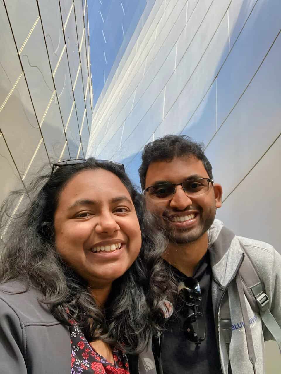 Shreeyeh and Kiran take a selfie in front of a modern building with reflective, angular metal panels under a clear blue sky. The sleek architectural design adds a contemporary and artistic vibe to the image.