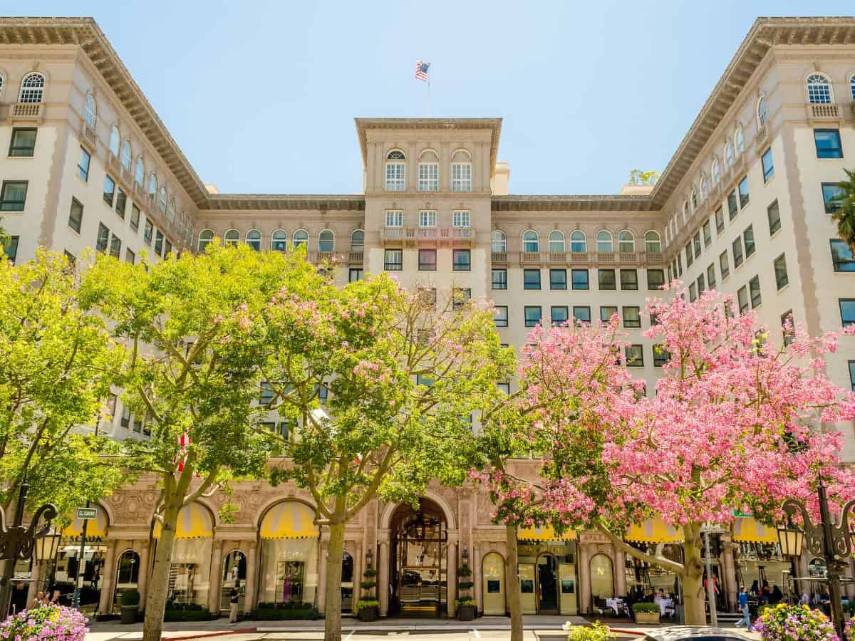 A daytime view of the Beverly Wilshire Hotel in Beverly Hills, showcasing its iconic Italian Renaissance architecture. The building is surrounded by lush trees and blooming pink flowers that line the front, creating a vibrant and inviting atmosphere.