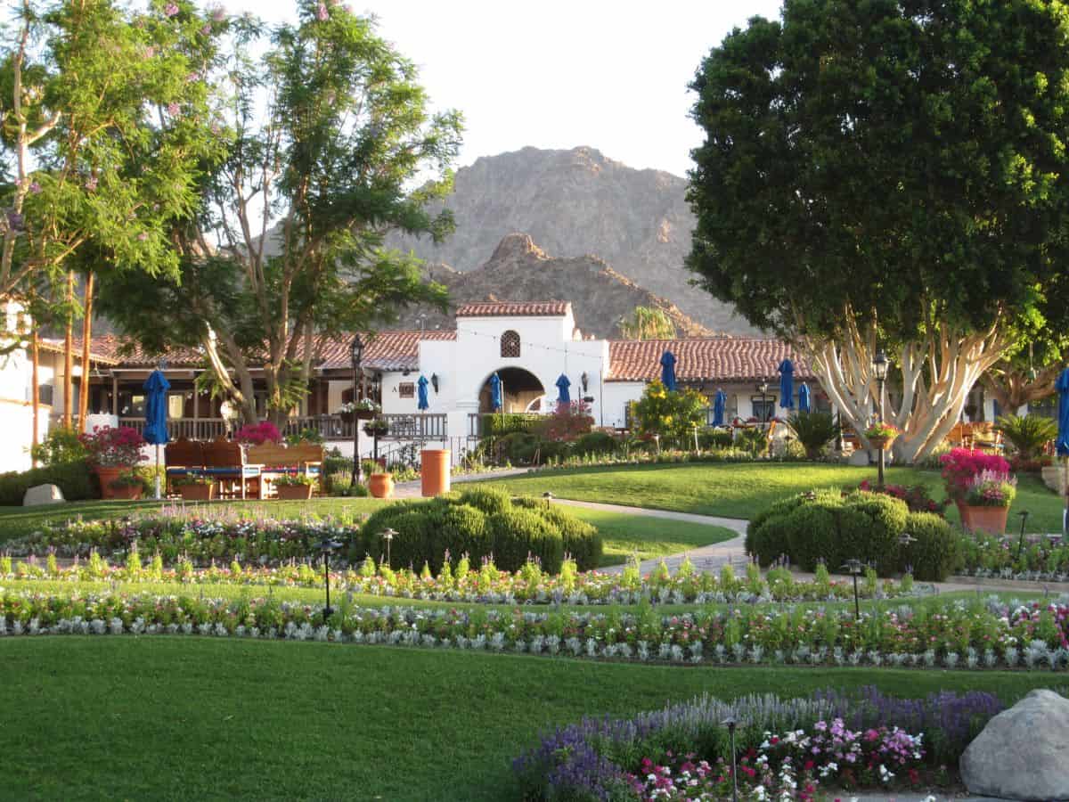 A peaceful daytime view of La Quinta Resort, showcasing its Spanish-style architecture. The lush green gardens are meticulously maintained with vibrant flower beds, and the surrounding mountains provide a serene backdrop.