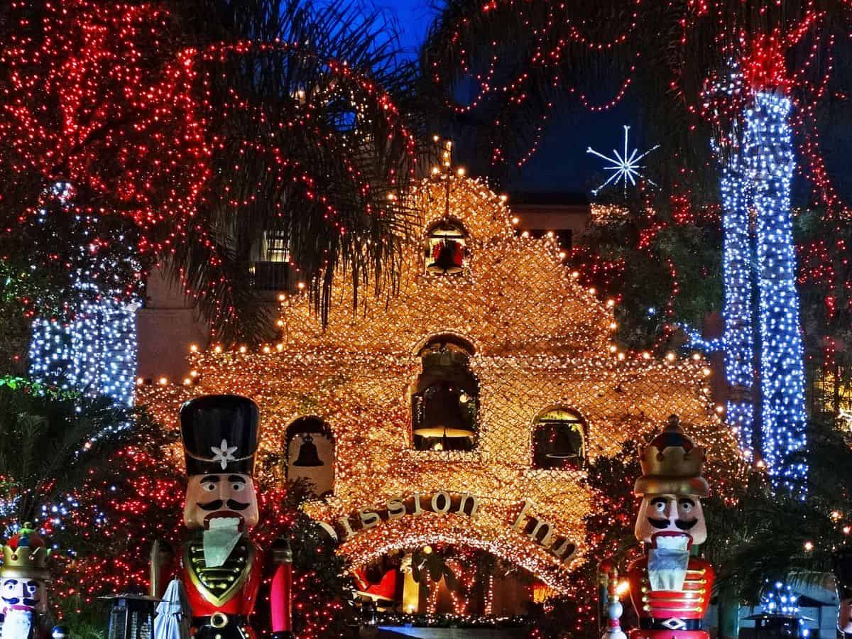 A dazzling nighttime view of the Mission Inn, decorated with thousands of sparkling red and white Christmas lights. Nutcracker statues stand at the entrance, adding to the festive display, while palm trees wrapped in lights complete the magical scene.