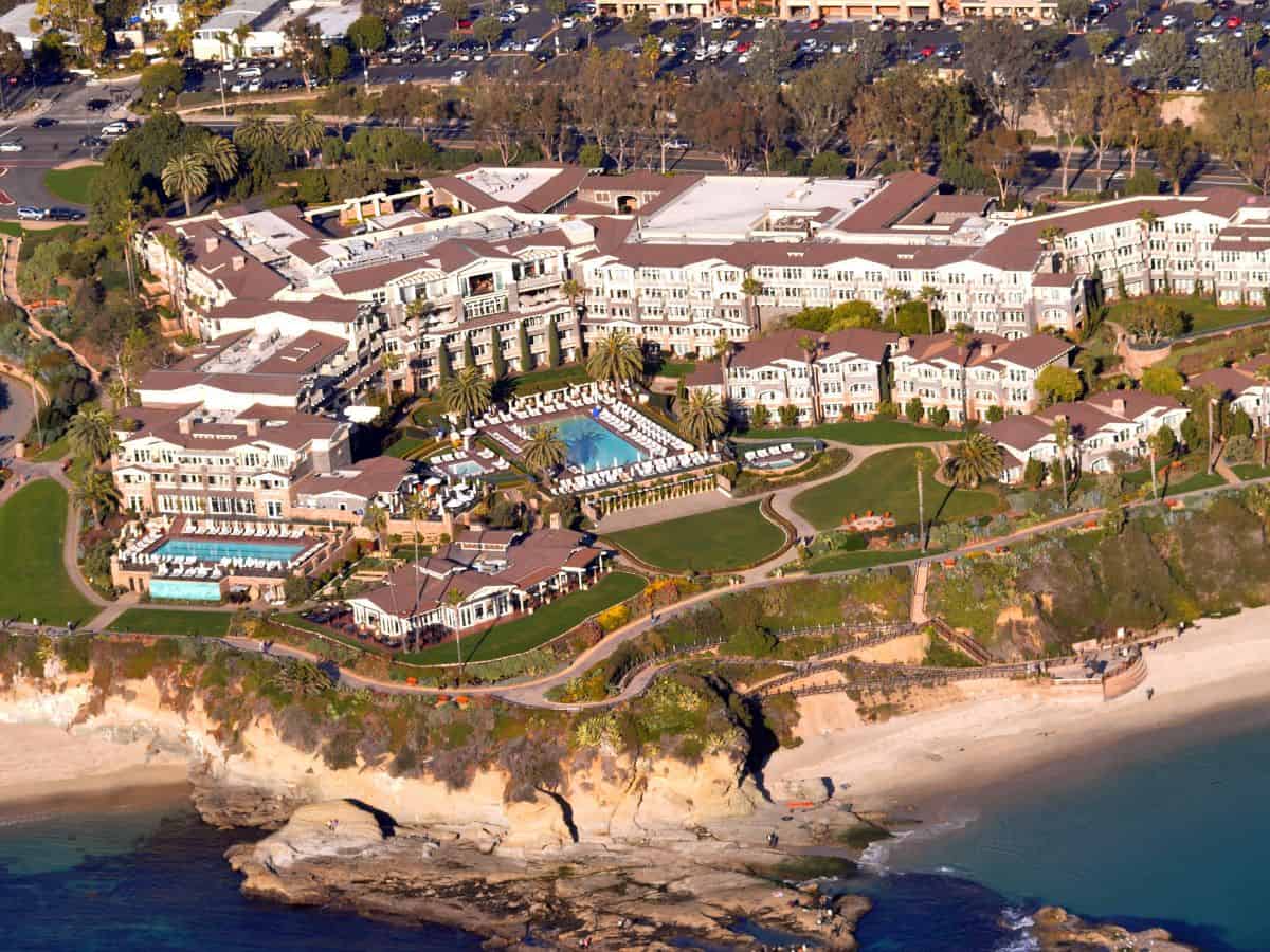 An aerial view of Montage Laguna Beach, capturing its luxurious resort layout. The property overlooks the Pacific Ocean, with multiple pools, well-manicured gardens, and a private beach, all bathed in natural sunlight.