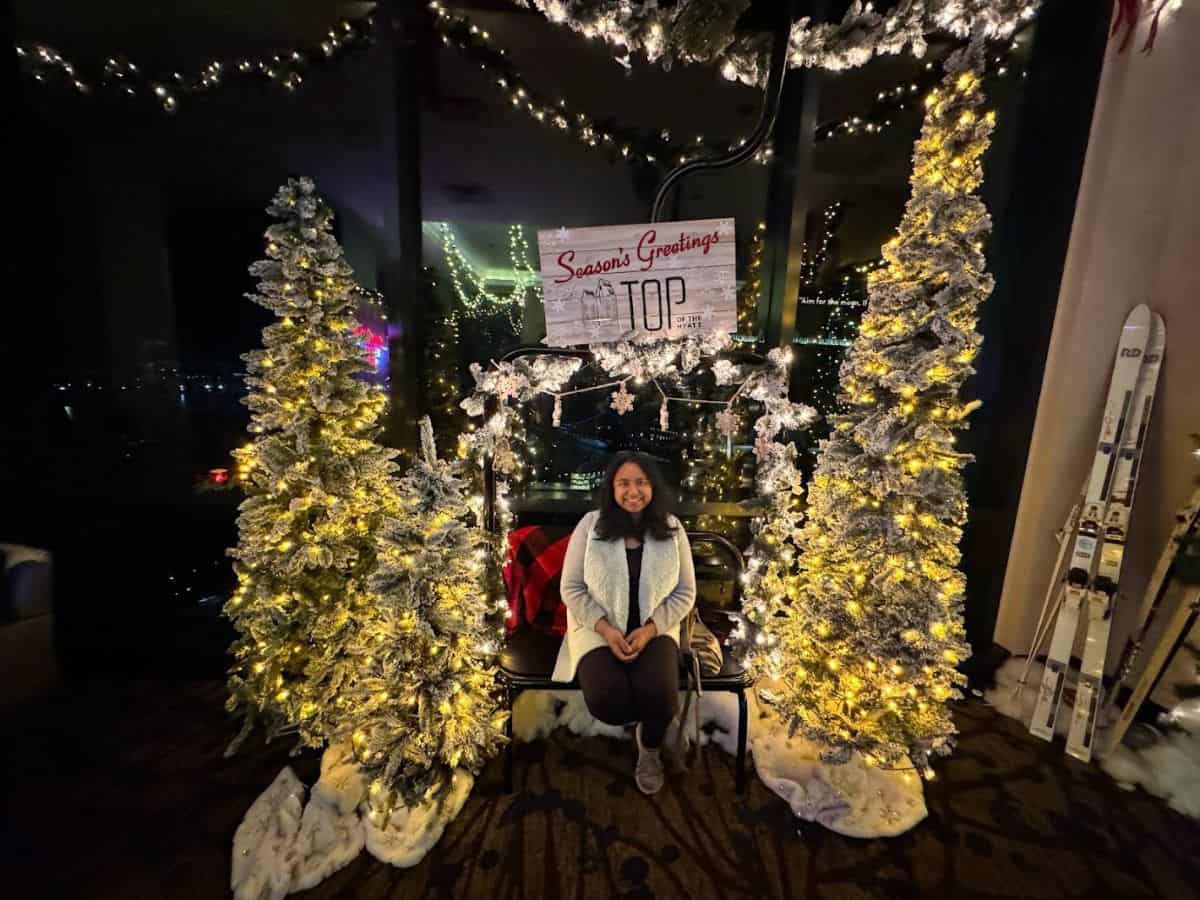 A cozy Christmas photo featuring Shreeyeh seated on a bench surrounded by festive decorations. Two tall, snow-dusted Christmas trees with glowing yellow lights frame the bench. Behind her is a festive sign reading "Season's Greetings Top" set against a backdrop of large glass windows revealing a nighttime city view with holiday lights in the distance.